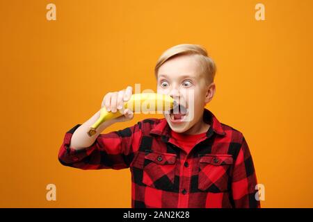 Le garçon mange des fruits, une alimentation saine pour les enfants. Beau garçon caucasien souriant dans la chemise rouge sur fond jaune. Horizontale, droite. Banque D'Images