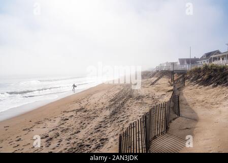 Lonely surfer carrying un surf le long du rivage d'une plage de sable sur un matin d'automne brumeux Banque D'Images