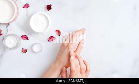 Concept de soins de la peau à la main. Vue de dessus les mains d'appliquer la crème hydratante bio, pots de crème cosmétique et pétales rose sur table de marbre. Natur Banque D'Images