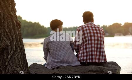 Adolescent mâle et son père assis sur la pierre près de lac et parler de la vie Banque D'Images