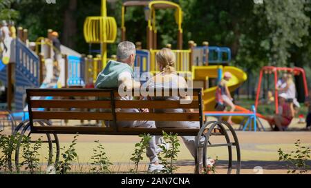 Couple assis sur un banc près de l'aire de jeux, regarder des petits-enfants jouant Banque D'Images