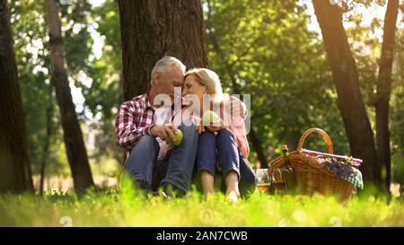 Extrêmement heureux vieux couple reposant sur l'herbe, tenant les pommes et l'embrassant, aire de pique-nique Banque D'Images