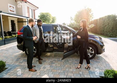 Jolie vendeuse de voiture en affaires aide à porter deux jeunes hommes d'multiethnical les clients à prendre la décision montrant une nouvelle voiture, l'ouverture des portes de voiture Banque D'Images