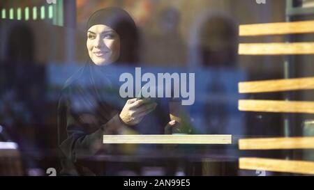 Belle dame musulmane holding coffee et smartphone dans les mains, assis dans cafe Banque D'Images