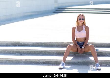 Mettre en place jeune fille avec de longs cheveux blonds dans une sports top, short et baskets se trouve sur l'escalier se reposant Banque D'Images