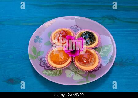 Pâte de barquettes rempli avec divers fruits confits, servi dans la plaque de verre rose sur table en bois bleu, décoré de fleurs roses Banque D'Images
