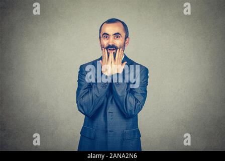 Surpris étonné d'affaires, man in suit holding ses mains sur ses joues, le visage. Course mixte modèle barbu isolé sur fond gris mur studio avec Banque D'Images