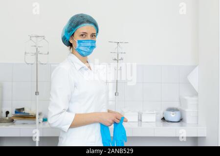 Une infirmière en blouse blanche met sur les gants avant une procédure médicale dans une chambre de traitement lumineux. Banque D'Images