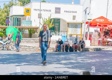Naxos Filoti - 13 août 2019 ; Middle aged homme grec avec de longs cheveux gris traverse rue où quatre touristes prennent une pause assis sur le pavé d'o Banque D'Images