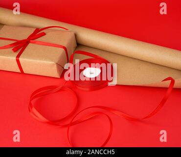 Rouleau de papier brun d'emballage, d'une bobine de ruban rouge et un cadeau enveloppé dans du papier kraft brun sur un fond rouge, faisant de surprises Banque D'Images