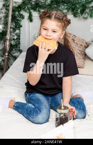 Peu cute belle fille est assise en tailleur sur un lit dans un jean bleu et une chemise noire de manger des pizzas et boire dans le contexte d'une chambre à la N Banque D'Images