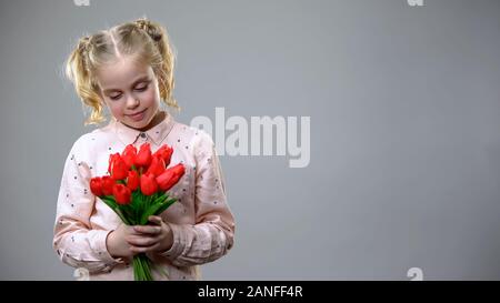 Adorable petite fille à la recherche sur les fleurs rouges dans les mains, fond gris, ambiance de vacances Banque D'Images