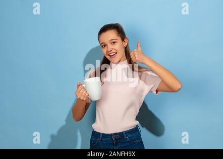 Café, thé, boissons montre pouce vers le haut. Caucasian girl's portrait sur fond bleu. Beau modèle dans des vêtements décontractés. Concept d'émotions humaines, expression du visage, ventes, ad. Copyspace. L'air heureux. Banque D'Images