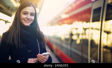 Jeune femme entrant sur smartphone sur près de la plate-forme et souriant pour appareil photo Banque D'Images