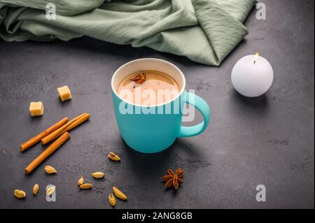 Thé traditionnel indien masala chai dans une tasse bleue. Bougie allumée, la cannelle, l'anis, la cardamome, le sucre à côté d'un fond texturé noir Banque D'Images