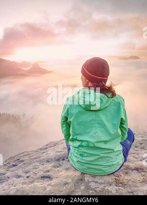 Belle femme de corps assis dans un windcheater vert sur le sommet de montagne regardant le lever du soleil sur une mer de brouillard Banque D'Images