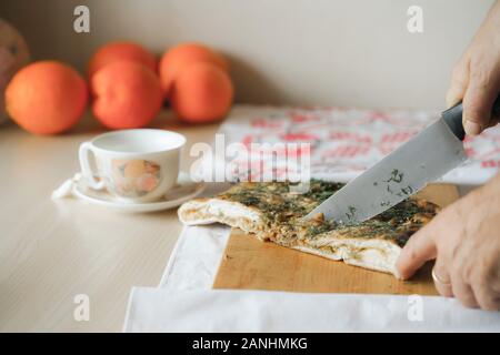 Man slicing up tourte aux légumes avec un couteau de cuisine sur la planche à découper. Sur l'arrière-plan sont orranges et tasse de thé. La nourriture végétarienne maison concept. Banque D'Images