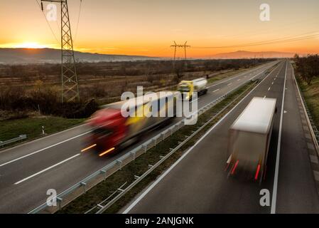 Camions et voitures de livraison en conduite à grande vitesse sur une route à travers le paysage rural. Mouvement flou rapide sur l'autoroute. Scène de fret sur le mois Banque D'Images