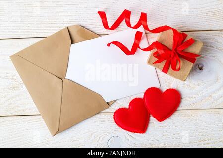 Carte de vœux vides dans une enveloppe brune, boîte-cadeau avec ruban arc et deux coeurs valentine rouge sur une surface en bois. Copier l'espace. L'amour et des sentiments concept Banque D'Images