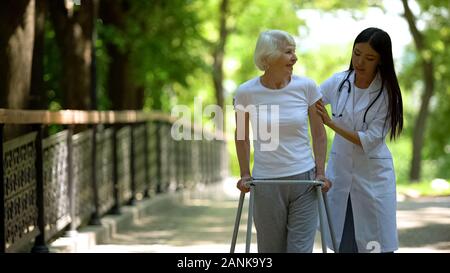 Caring nurse helping senior femme handicapée avec cadre walk in park, rehab Banque D'Images