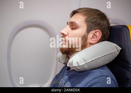 Jeune homme européen séduisant de 30 ans avec une barbe dormant, reposant avec un oreiller de cou gonflable dans un avion. Confort, stress dans un avion Banque D'Images