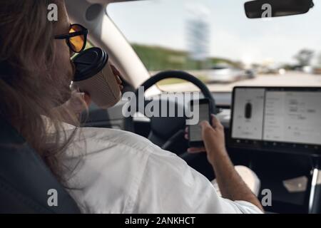 Mode de transport. L'homme aux cheveux longs dans les lunettes de voyager assis à l'intérieur de la voiture électrique la conduite sur la consommation de café de pilote automatique du smartphone navigation Banque D'Images