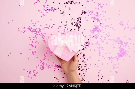 Femme main tenant une boîte en carton rose avec un arc sur un fond rose avec des confettis multicolores, concept de donner un cadeau pour des vacances Banque D'Images