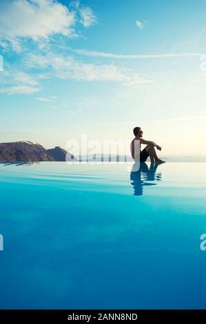 L'homme assis sur le bord d'une piscine à débordement surplombant la Méditerranée pittoresque vue sur la caldeira de Santorin, Grèce Banque D'Images