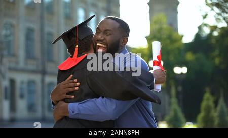 Cheerful père et fils diplômés hugging extérieur, réalisation de l'étude, de l'éducation