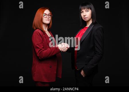 Stock photo portrait de deux femmes d'affaires réussie dans l'élégant convient à serrer la main de faire un deal. Looking at camera, isoler sur noir. Banque D'Images