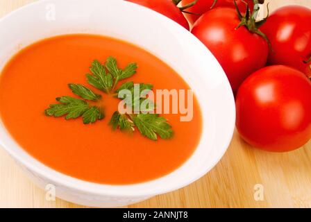 Bol de soupe aux tomates avec des feuilles de persil plat italien pour une saisie-arrêt. Un tas de vigne tomates mûres avec des tiges sont en arrière-plan. Banque D'Images