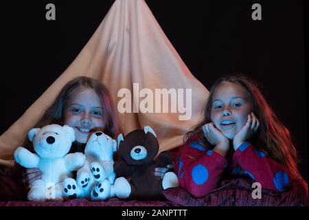 Les enfants et les temps d'amusement concept. PJs partie pour les enfants. Les filles avec des visages heureux de jouer avec fantaisie. Kids wearing red pyjama au lit sur fond noir. Les amis s'amusant avec des jouets dans une tente. Banque D'Images