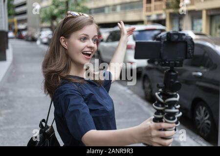 Jeune fille caucasienne souriante femme faisant un blog vidéo (vlog) avec caméra à l'extérieur dans la rue Banque D'Images