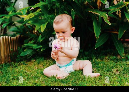 Bébé en couche sans vêtements assis sur l'herbe de grignoter une fleur. Banque D'Images