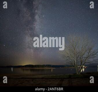 Voie lactée d'un arc sur un arbre sans feuilles sur la rive du lac Jindabyne avec la ville de Jindabyne en arrière-plan Banque D'Images