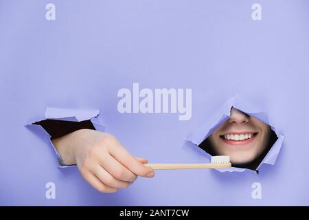 Femme positive avec dents agréable sourire sur le visage, la tête et le bambou eco permet grâce à la brosse à dents dans le trou déchiré papier violet, copie de l'espace pour votre promotion Banque D'Images