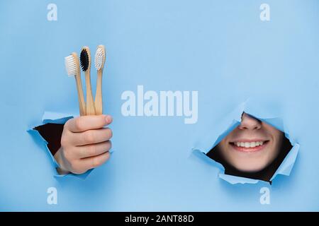 Femme positive avec dents agréable sourire sur le visage, la tête et le bambou eco permet de brosses à dents à travers le trou déchiré en papier bleu, copie de l'espace pour votre promotion Banque D'Images