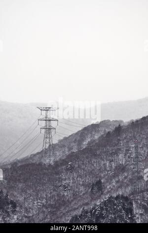 Un pylon électrique (tour de transmission, tour de puissance) se dresse au sommet d'une montagne enneigée et couverte d'arbres dans la campagne du Japon par un jour nuageux. Banque D'Images