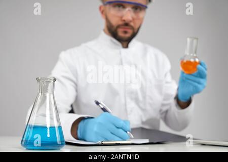 Avant récolte de jeune chimiste en blouse de laboratoire blanche, casque et gants assis au bureau, tenant une fiole orange, regardant l'appareil photo et écrire dans un dossier. Mise au point sélective de la fiole avec liquide bleu sur la table. Banque D'Images