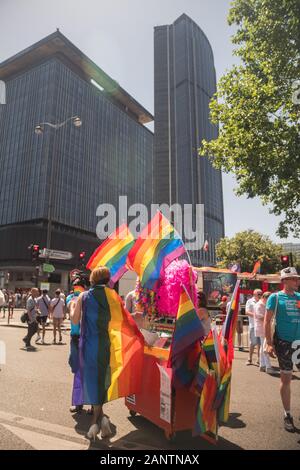 29 Juin 2019, Paris, France. Gay Pride Parade Day. Travestite devant un stand de goodies (drapeaux arc-en-ciel). Le skyskraper 'la Tour Montparnasse'. Banque D'Images