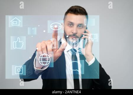 Vue de face de jeune homme d'affaires en costume noir parlant au téléphone au bureau. Mise au point sélective de l'écran des cartes tactiles numériques, homme barbu en cliquant sur le bouton virtuel. Concept de gadgets, numérisation. Banque D'Images