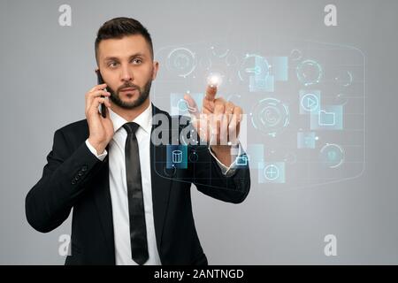 Vue de face de jeune homme d'affaires en costume noir parlant au téléphone au bureau. Homme barbu sérieux en cliquant sur le bouton virtuel sur l'écran des cartes tactiles numériques. Concept de gadgets, numérisation. Banque D'Images