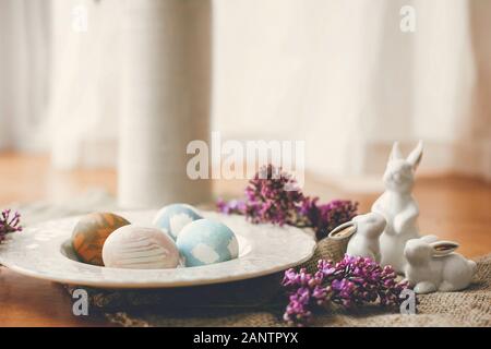 Les oeufs de Pâques sur élégant vintage plate, petits lapins blancs et lilas fleurs sur le tissu sur une table en bois rustique. Composition de pâques Rural teint naturel Banque D'Images