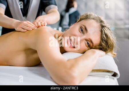 Jeune blonde curly woman receiving massage dos manuel classique dans medical spa centre, looking at camera et souriant. Vue de côté Banque D'Images