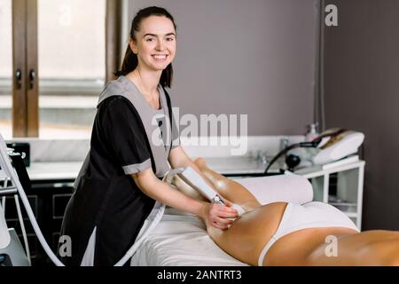 Young smiling woman cosmetologist avec rouleau faire massage des fesses pour sa cliente dans un spa moderne centre médical. Banque D'Images