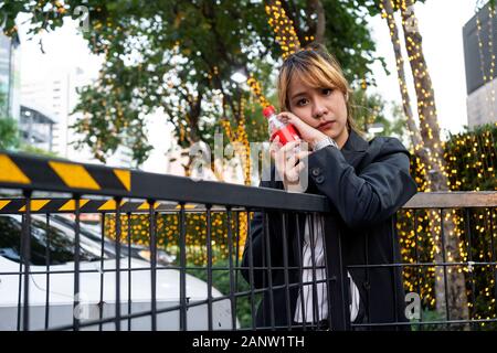 La moitié des cheveux blonds attachés femme en noir et blanc vêtements hug la bouteille de boisson gazeuse à la clôture en parking avec décoration lumineuse. Banque D'Images