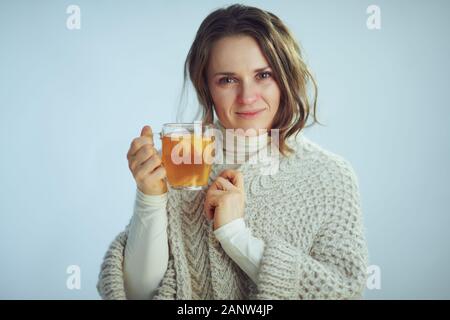 Portrait de malade de 40 ans femme au foyer moderne en rouleaux et du cou cardigan montrant tasse de thé au gingembre, citron et miel isolé sur winte Banque D'Images