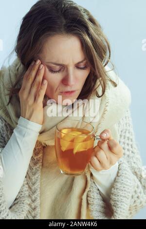 Mauvais femme moderne en rouleaux et du cou cardigan holding tasse de thé au gingembre, citron et miel et avoir des maux de tête contre la lumière d'hiver b bleu Banque D'Images