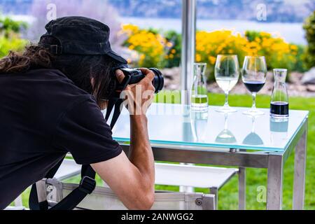 Photographe l'homme aux cheveux longs vêtu de noir vue arrière arrière alors qu'il prend des photos de verres à vin avec un appareil photo reflex numérique professionnel, vin winery concept Banque D'Images