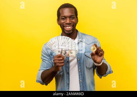 Portrait d'homme heureux en denim chemise décontractée holding dollars et bitcoin physique, smiling at camera, sentiment de succès et riche avec cryptocur Banque D'Images